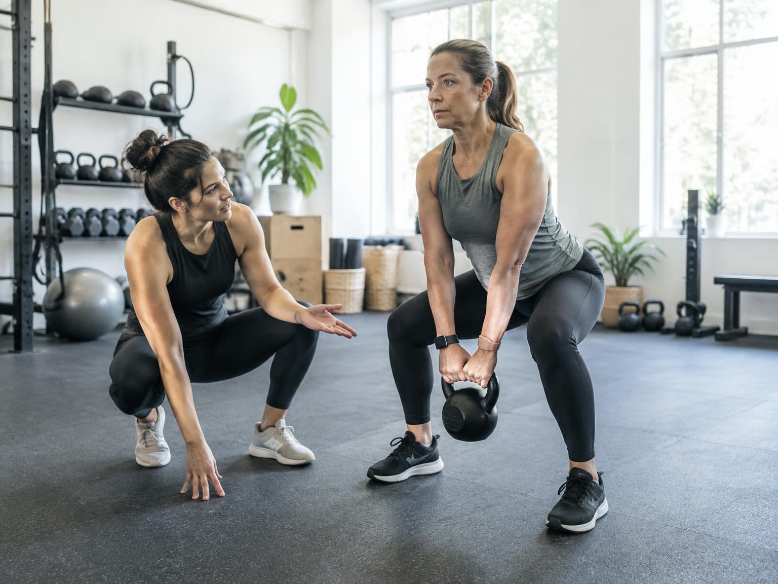 A personal trainer coaching a member through a kettlebell exercise in a bright studio