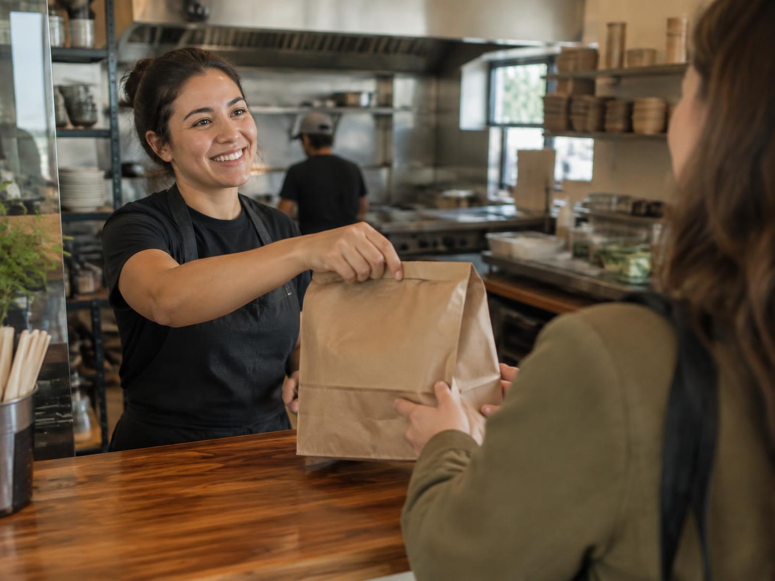 A counter worker handing a paper bag of food to a customer in a small restaurant